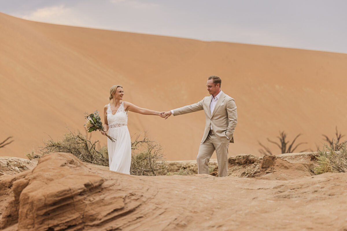 Bride and groom walking hand in hand across dunes of Sossusvlei during their wedding photoshoot, image by Mariette du Toit Photography