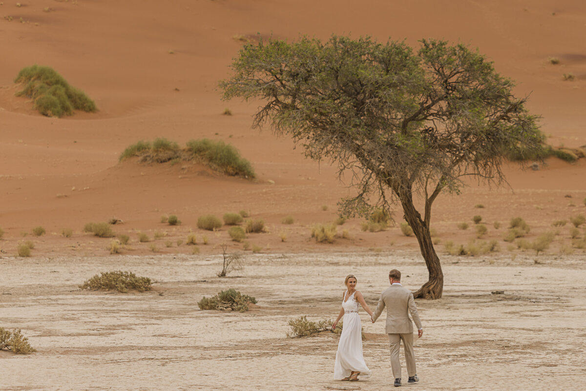 Bride and groom walking hand in hand across the white clay pan of Sossusvlei during their wedding photoshoot, image by Mariette du Toit Photography