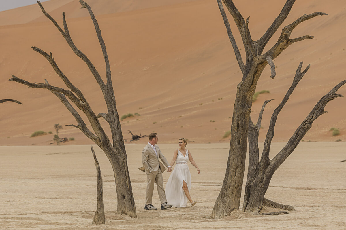 Couple standing in Deadvlei, framed by ancient camel thorn trees and soft desert hues, photographed on their Namibian honeymoon, image by Mariette du Toit Photography