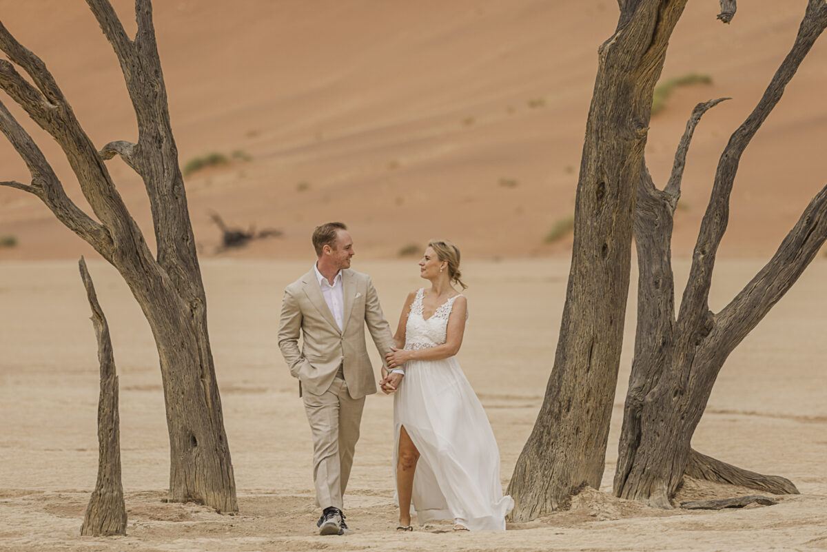 Couple walking hand-in-hand in Deadvlei, framed by ancient camel thorn trees and soft desert hues, photographed on their Namibian honeymoon, image by Mariette du Toit Photography