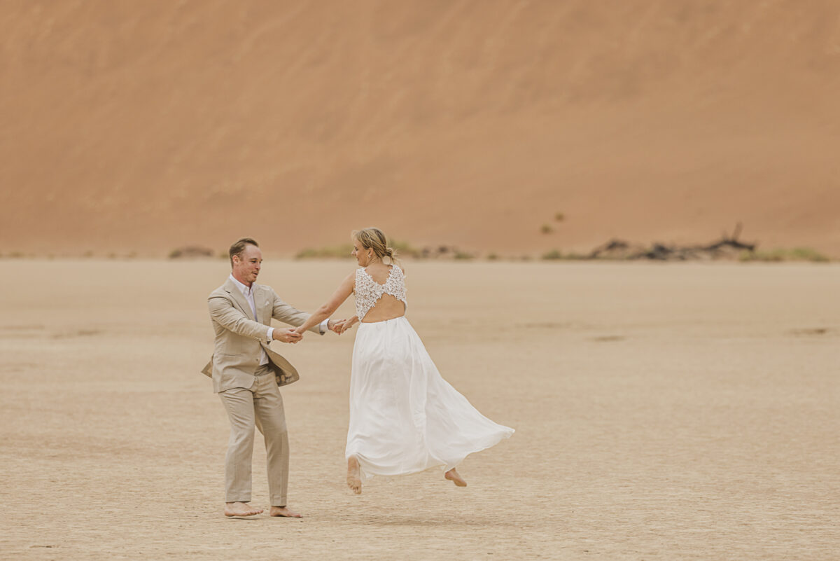Bride and groom walking hand in hand across the white clay pan of Sossusvlei during their wedding photoshoot, image by Mariette du Toit Photography