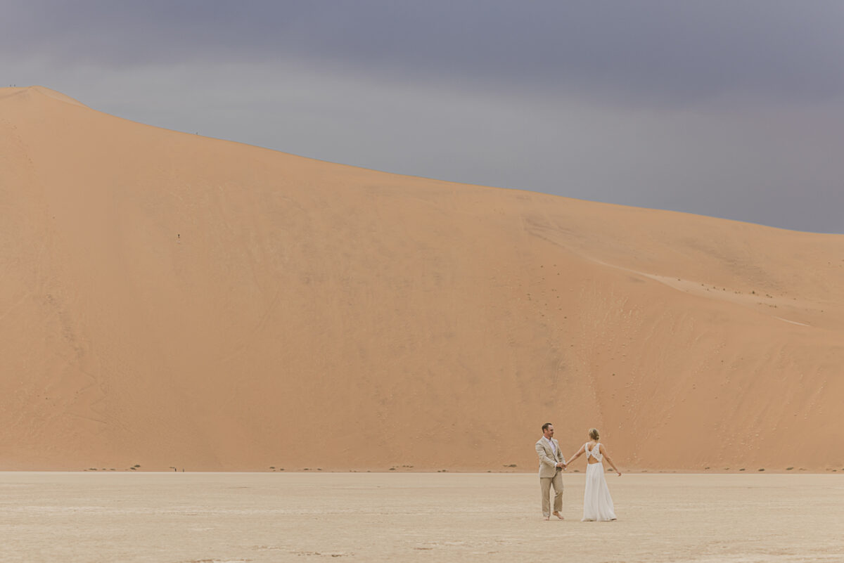 Bride and groom walking hand in hand across the white clay pan of Sossusvlei during their wedding photoshoot, image by Mariette du Toit Photography