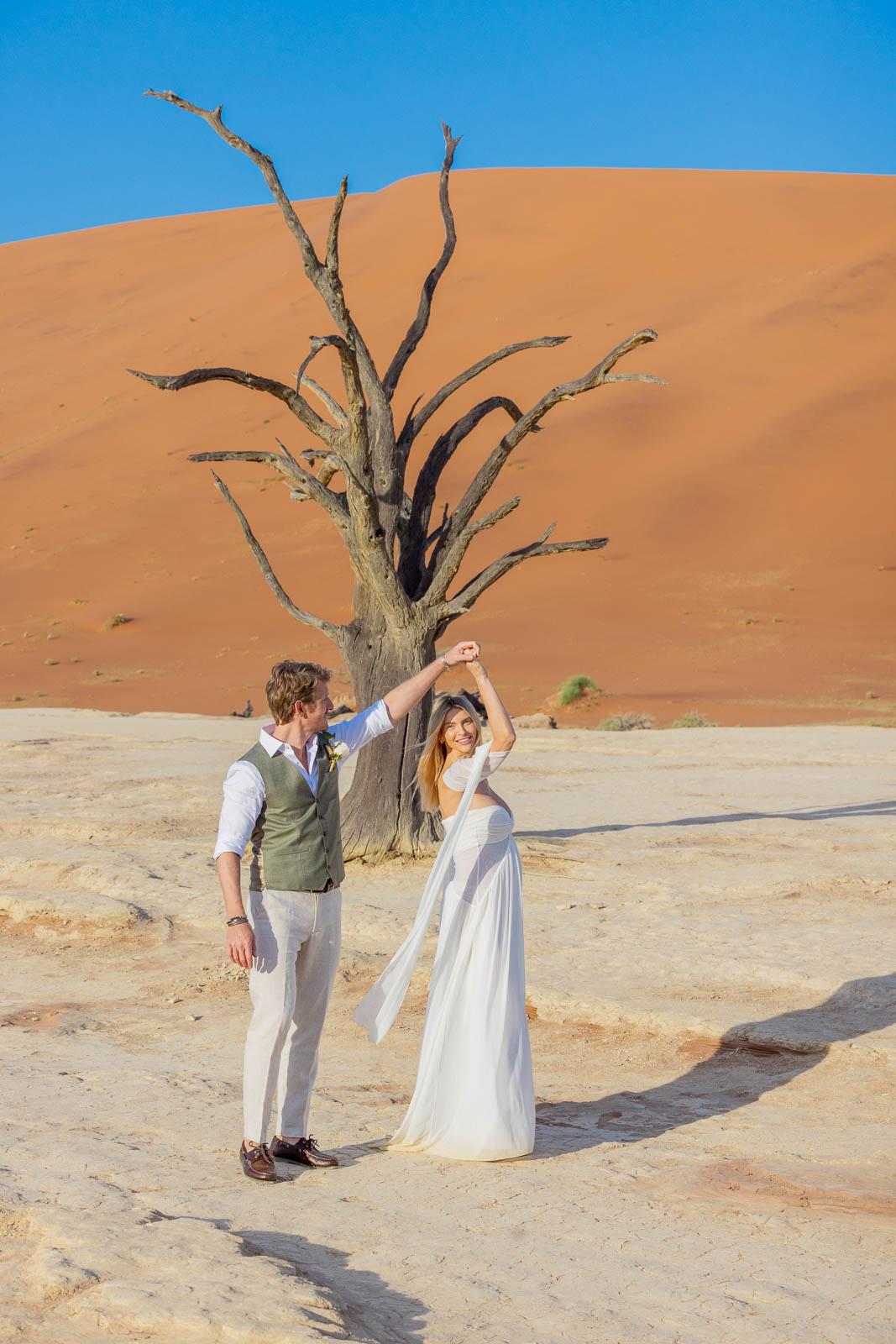 Newlyweds celebrating with arms raised beneath a dead tree and red dune in Deadvlei